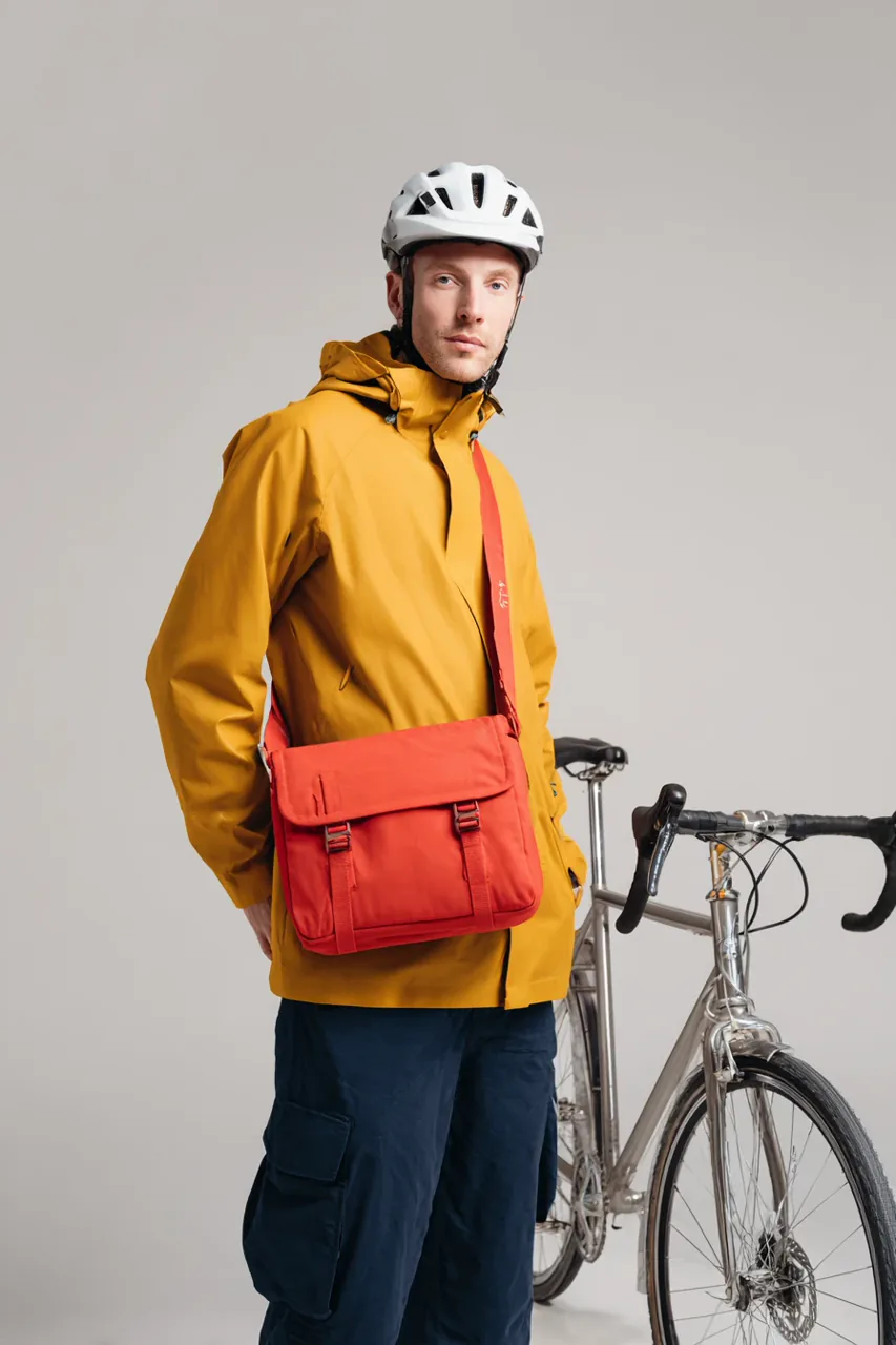 Bicycle courier in a mustard rain jacket with a red messenger satchel beside a partially visible silver bicycle